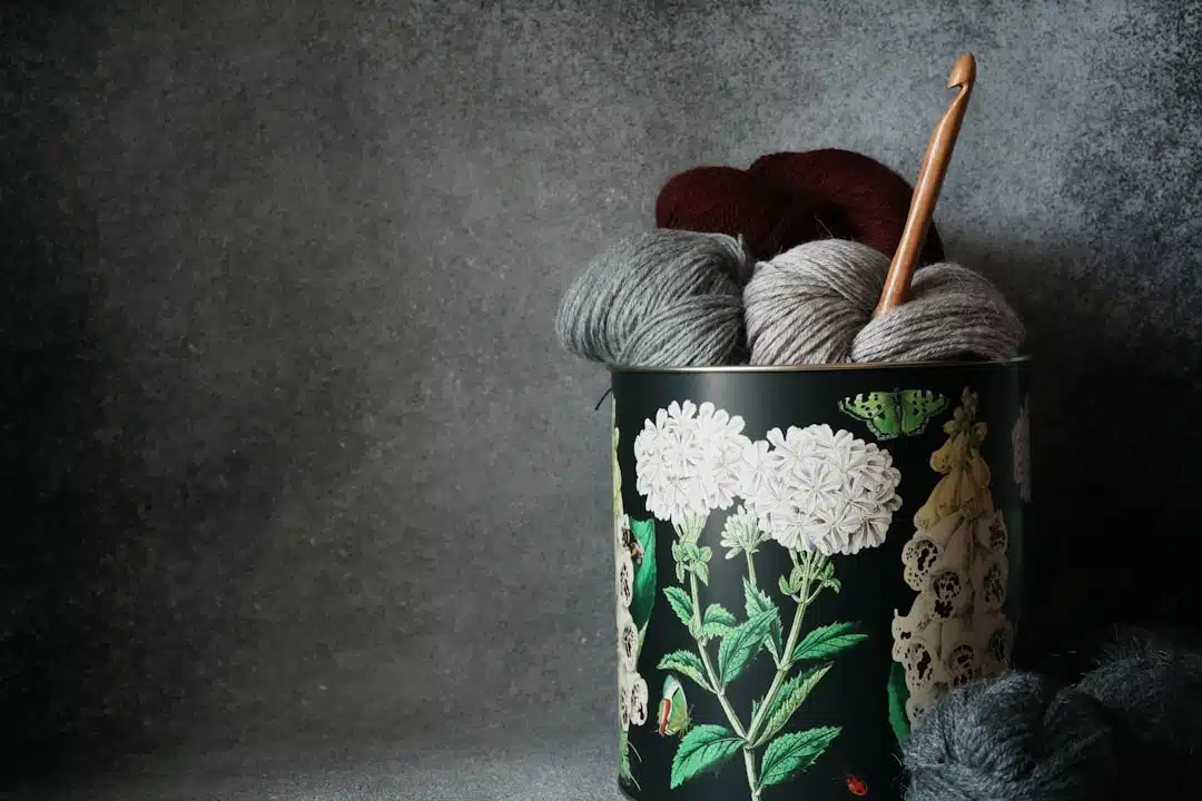 A tin with floral design holds yarn balls and a crochet hook, with gray yarn on a dark background.