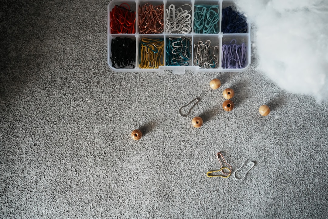 A box of colorful paper clips, wooden beads, and hooks on a gray carpeted floor, with white fluff in the corner.
