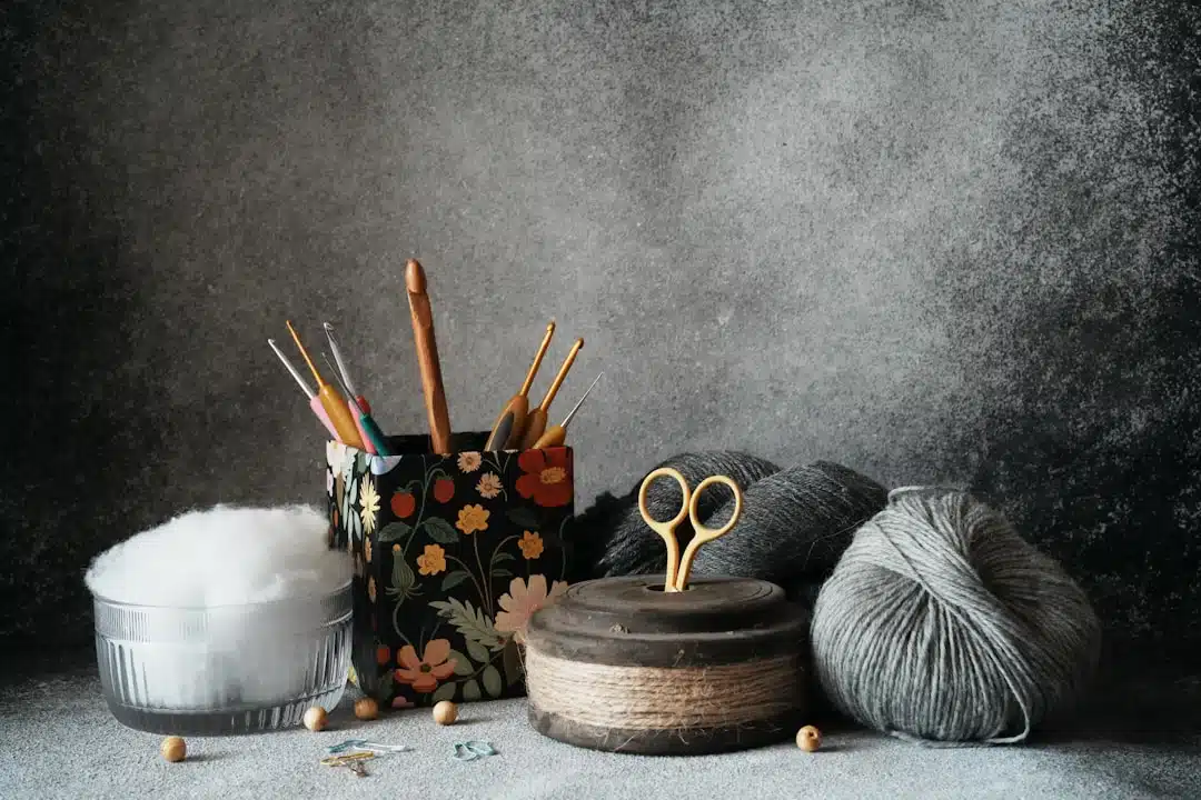 A floral cup holds crochet hooks beside yarn balls, scissors, stuffing, and wooden beads on a gray surface.