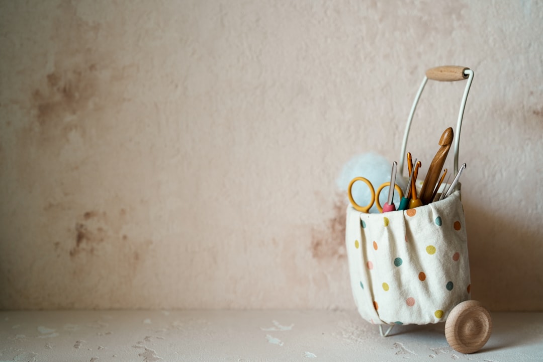 A polka dot fabric basket on wheels holds craft supplies like scissors, crochet hooks, and yarn against a beige wall.