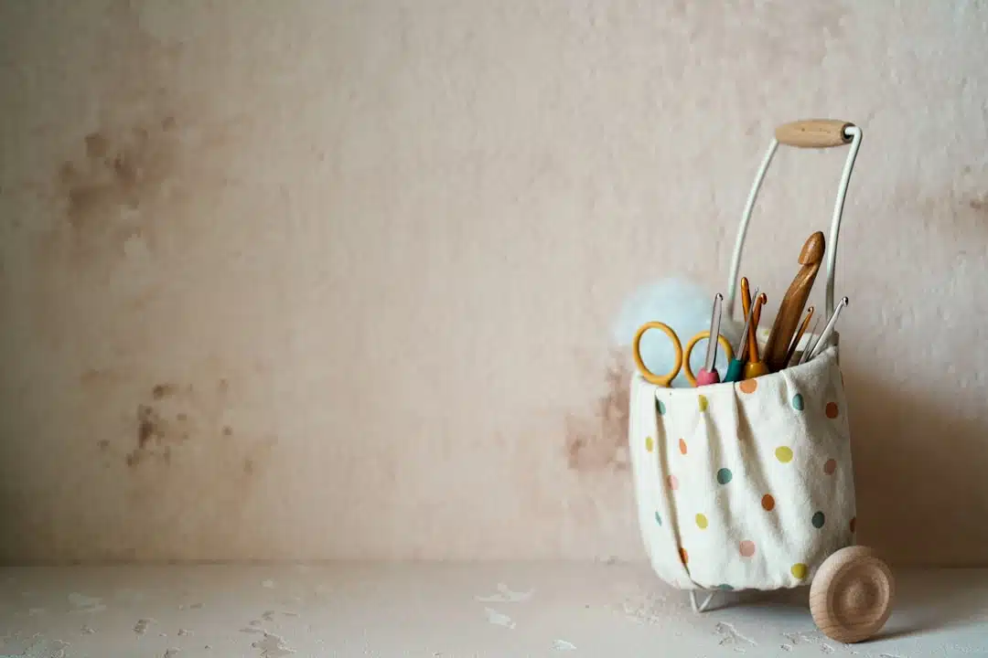 A polka dot fabric basket on wheels holds craft supplies like scissors, crochet hooks, and yarn against a beige wall.