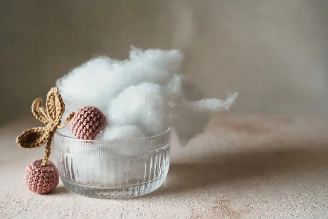 Glass bowl with white cotton-like stuffing, crocheted pink cherries, and a crocheted brown leaf on a beige surface.