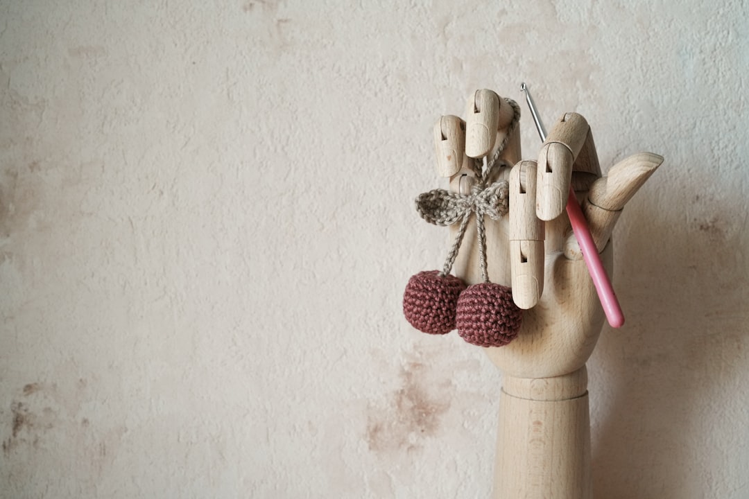 A wooden mannequin hand holds crocheted cherries and a pink-handled crochet hook against a beige wall.