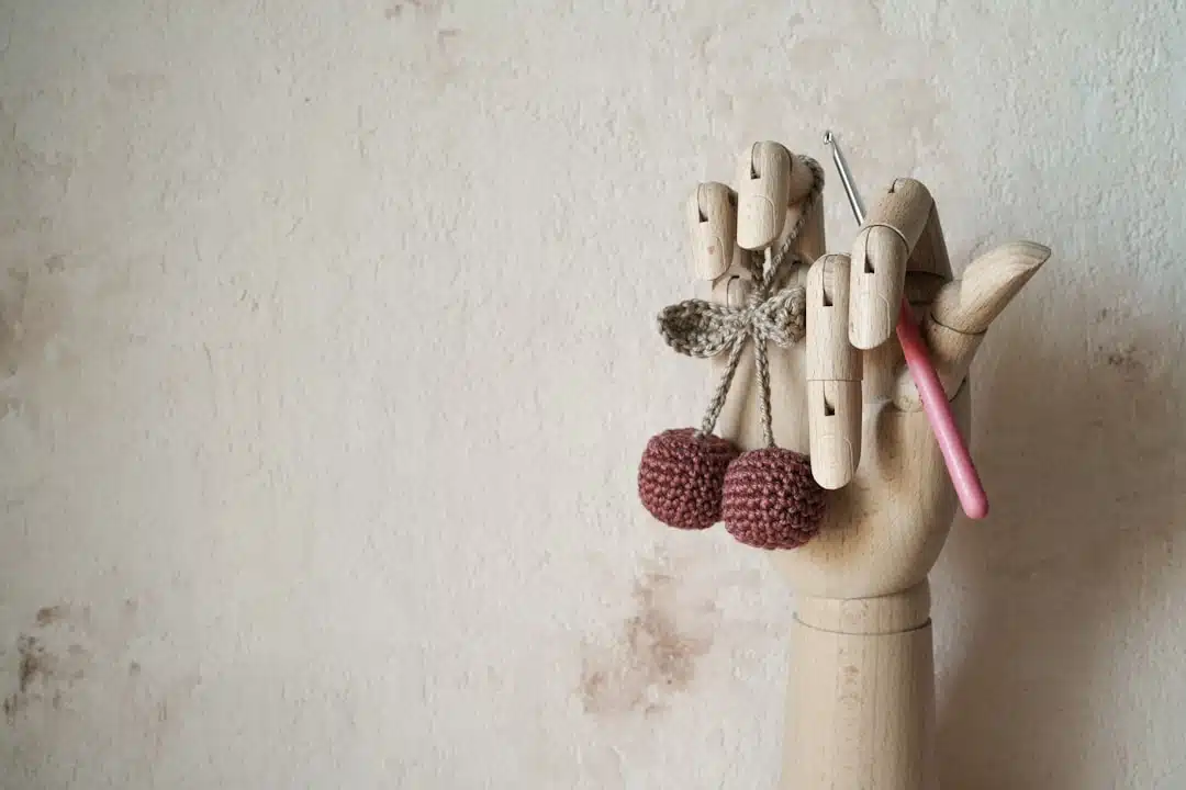 A wooden mannequin hand holds crocheted cherries and a pink-handled crochet hook against a beige wall.