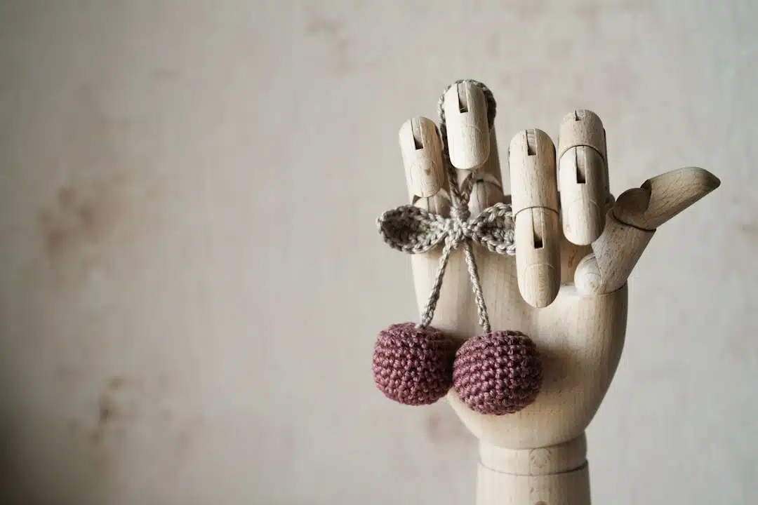 A wooden mannequin hand displays two crocheted cherries tied with a string against a neutral background.