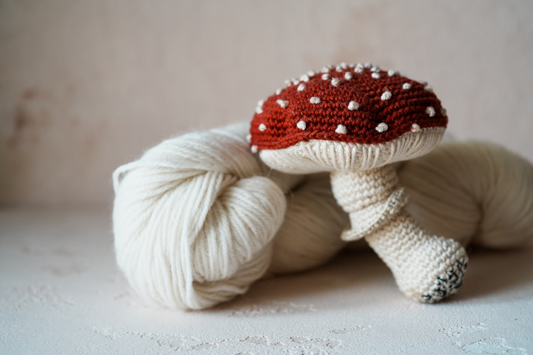 A crocheted red and white mushroom rests beside a skein of cream-colored yarn on a light surface.