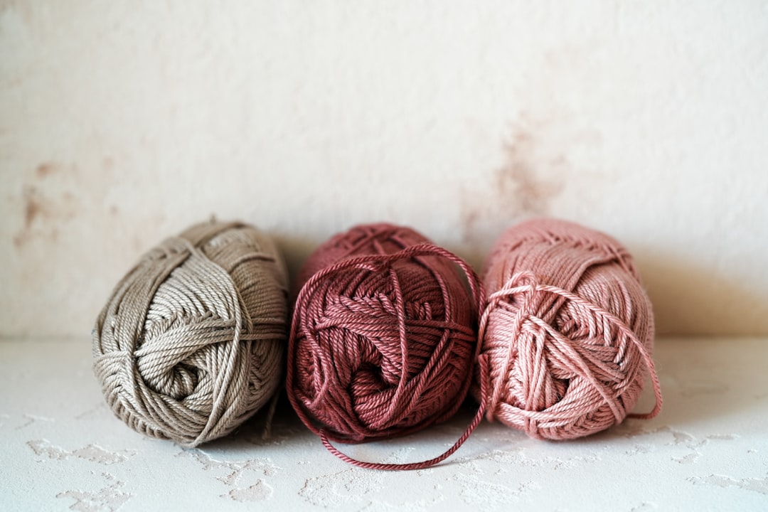 Three balls of yarn in beige, maroon, and pink colors are placed side by side on a light textured surface.