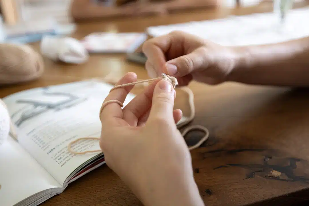 Hands holding yarn and crocheting with an open instruction book on a wooden table in the background.