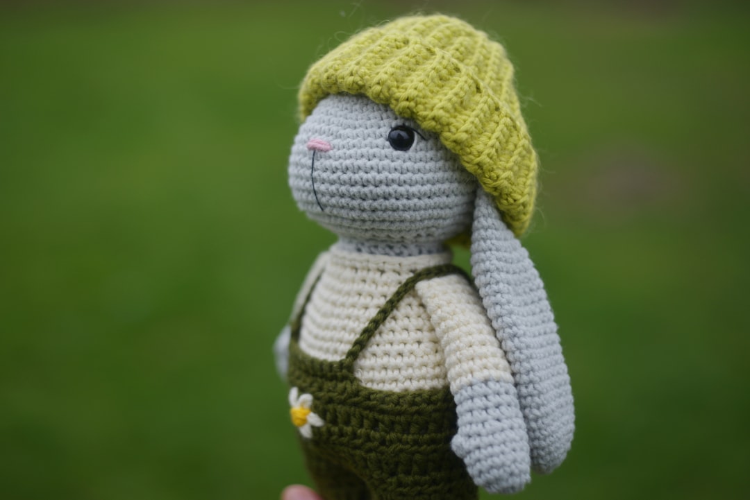 A crocheted bunny in green overalls and a yellow hat is held outdoors with green grass in the background.