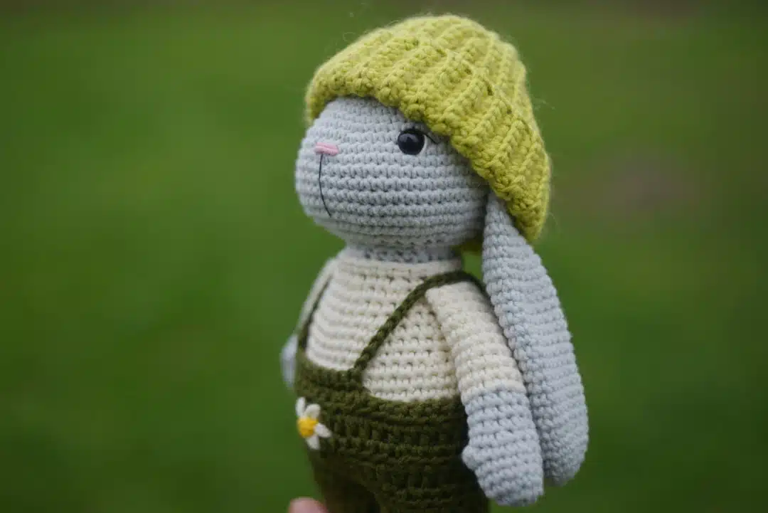A crocheted bunny in green overalls and a yellow hat is held outdoors with green grass in the background.