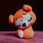 A small crocheted orange animal with closed eyes and white accents sits on a pink surface.