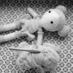 A crocheted doll lies next to a ball of yarn and a crochet hook on a patterned surface.