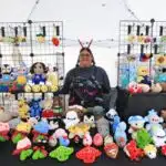 A person stands behind a booth displaying many colorful crocheted plush toys and keychains at a market.