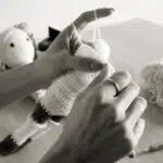 Hands crocheting a stuffed animal, with yarn, a hook, and finished toys on a table in black and white.