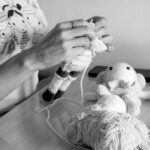 A person crochets a doll at a table with yarn, a finished toy, and other crafting supplies nearby.