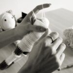 Hands crocheting a stuffed animal with yarn on a table; a finished toy and crochet hook are nearby.