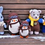 Five crocheted animal dolls, including a rabbit, penguins, a bear, and a cow, sit on a snowy wooden bench.