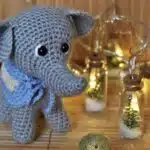 A crocheted gray elephant with a blue scarf stands by fairy lights and tiny tree jars on a wooden surface.