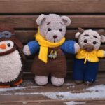Three plush toys—a penguin, a bear, and a cow—sit on a wooden bench with some snow on the seat.