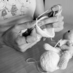A person crochets a white yarn piece next to a crocheted stuffed animal on a wooden table.