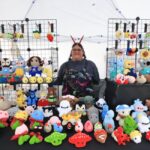 Person behind a table displaying colorful crocheted stuffed animals and characters at a market booth.