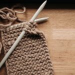 Brown yarn and two white knitting needles forming a piece of knitted fabric on a wooden table.