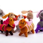 Seven colorful crocheted dachshund dog toys are grouped together on a white background.