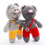 Two crocheted cats with pink noses, one in yellow pants and one in red pants, stand side by side.