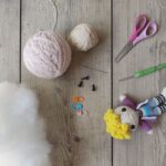 Yarn, scissors, crochet hook, stuffing, markers, doll eyes, and a crocheted doll on a wooden surface.