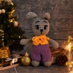 Crocheted gray bunny in purple overalls stands by a decorated Christmas tree with festive lights and gifts.