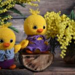 Two crocheted yellow chicks in clothes sit on logs, with mimosa flowers and greenery in the background.