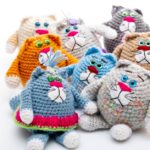 Eight colorful crocheted cat dolls with big eyes and pink noses arranged on a white background.