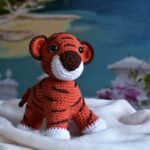 A crocheted tiger plush sits on a white blanket with a scenic seaside backdrop.