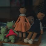Several crocheted dolls, including two girls and two frogs, are displayed on a table with a blurred background.